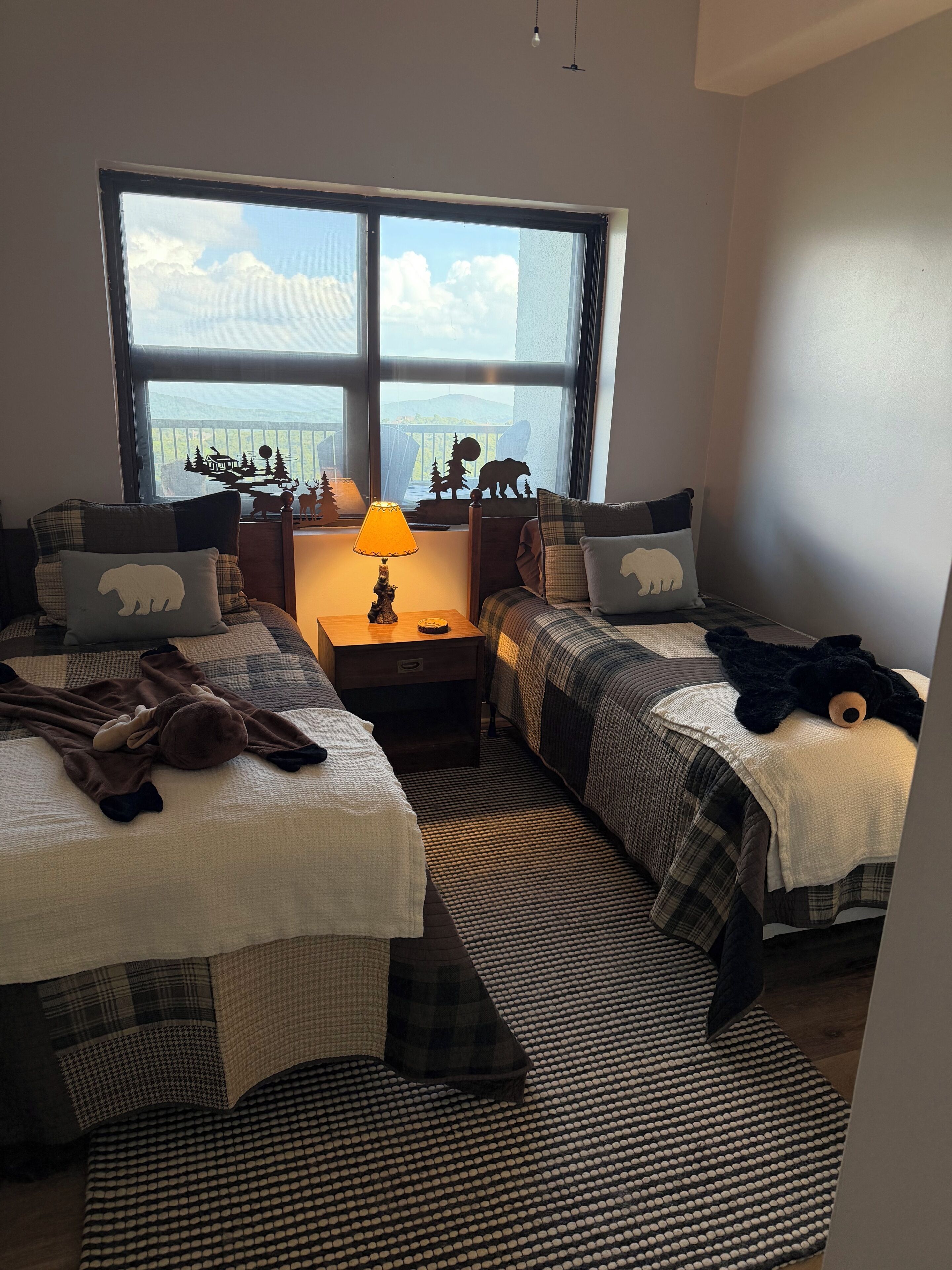 Une chambre avec deux lits, une fenêtre donnant sur un beau paysage, une lampe et un ours en peluche sur l’un des lits.