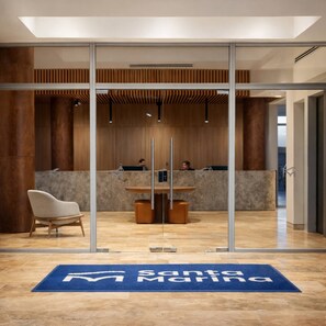 A reception area with a glass partition, a reception desk, a seating area with a chair, and a blue carpet with 'Santa Marina' text. - Ocean View 14th Floor Pools & Beach (Santa Marta)