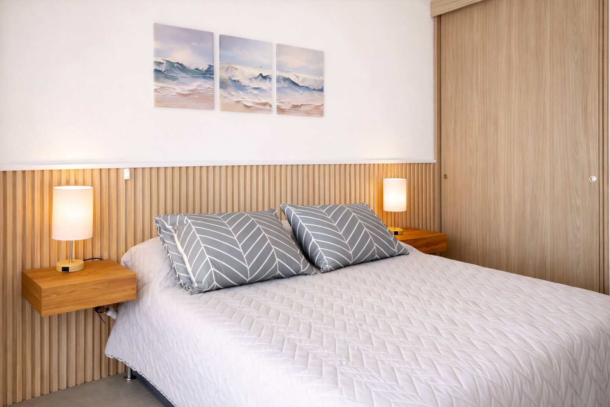Un lit bien fait, recouvert d’une couette blanche matelassée, avec deux oreillers à motifs géométriques et deux lampes de chevet posées sur des tables de nuit en bois.