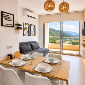A modern dining area with a wooden table set for two, a grey sofa, and a large window offering a view of a green landscape. - Ocean View 14th Floor Pools & Beach (Santa Marta)