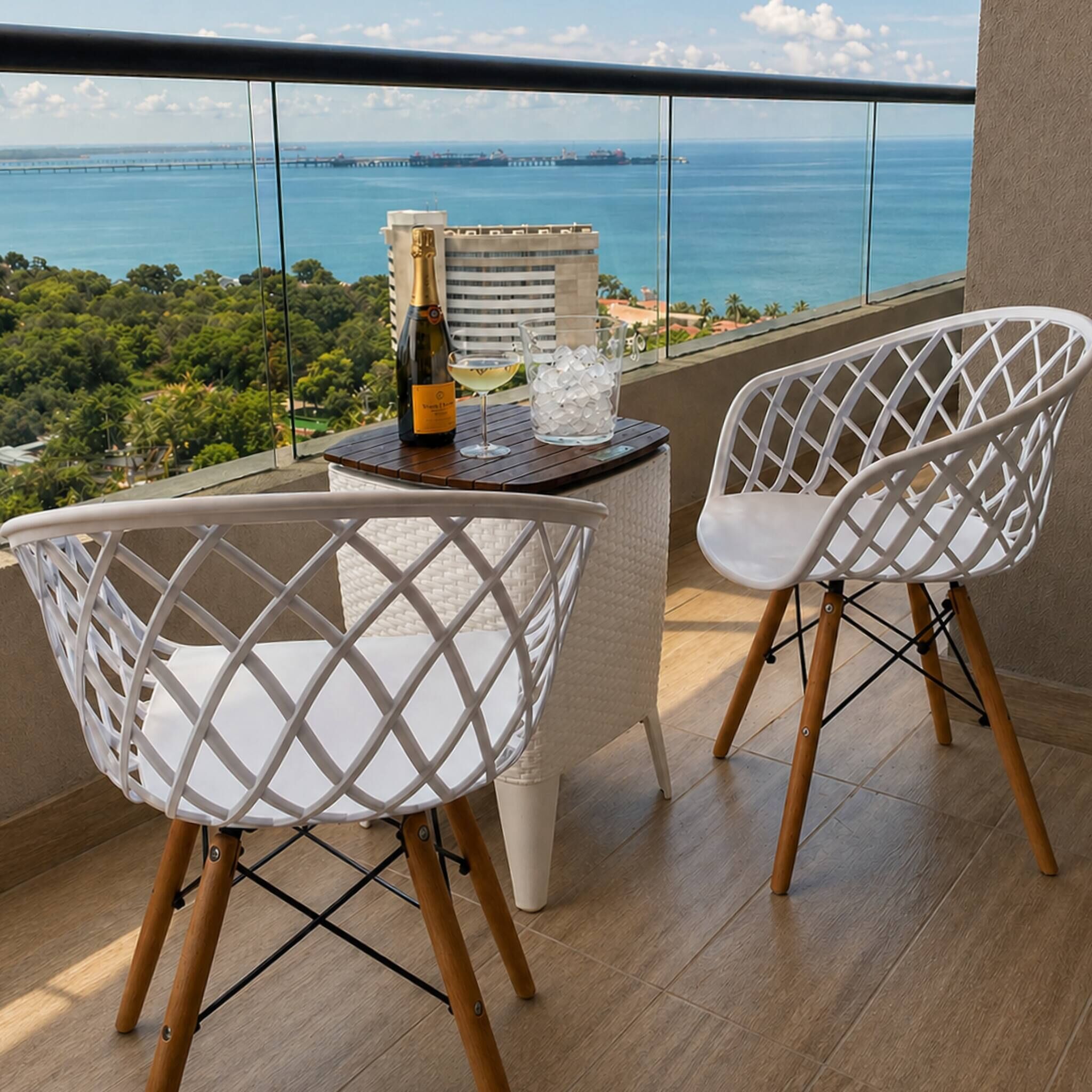 A balcony with two white chairs, a table with a bottle and glasses, and a view of the ocean and a bridge.