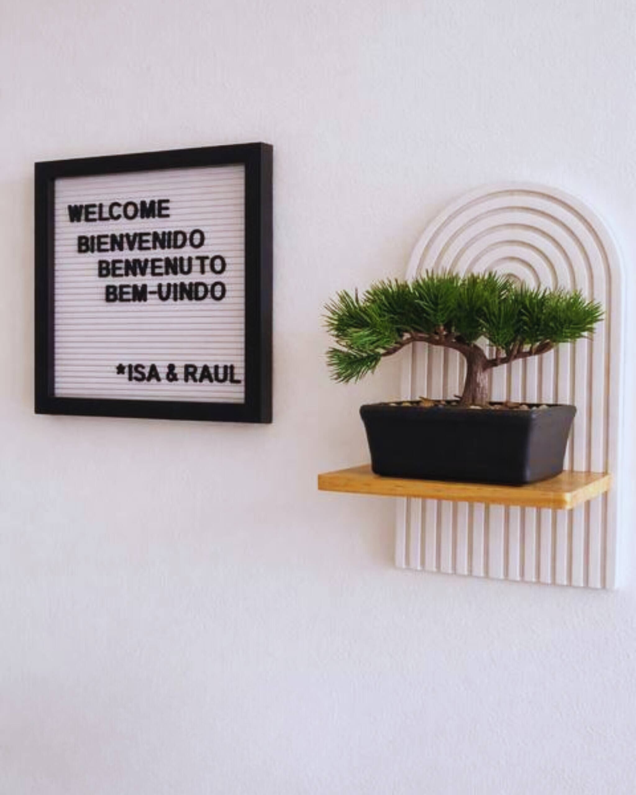 A framed welcome sign with multilingual greetings and a bonsai tree on a shelf.