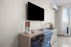 Ein modernes Hotelzimmer mit einem wandmontierten Flachbildfernseher, einem Holztisch mit Stuhl und einem großen Fenster mit Vorhängen.