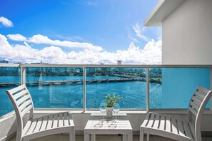 Ein Balkon mit zwei weißen Stühlen, einem kleinen Tisch mit einer Vase und zwei Weingläsern sowie Blick auf ein Gewässer mit einer Brücke und Gebäuden im Hintergrund.