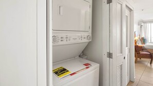 Un cuarto de lavado con lavadora y secadora de carga frontal, una puerta a otra habitación y una silla al fondo.