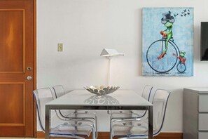 Een eetgedeelte met een glazen tafel, transparante stoelen, een witte muur, een schilderij van een kikker op een fiets, en een witte vloerlamp.