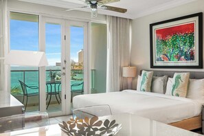 Ein Hotelzimmer mit einem Bett, einem Schreibtisch, einem Stuhl, einem Balkon mit Meerblick und einem Bild an der Wand.