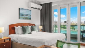 Hotelkamer met een bed, nachtkastje, stoel, tafel, airconditioning, schilderij en een glazen schuifdeur naar een balkon met uitzicht op de stad.