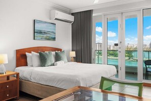 A hotel room with a bed, a nightstand, a chair, a table, an air conditioner, a painting, and a sliding glass door to a balcony with a city view. - Lagoon Front 3BR - Balconies & Pool (San Juan)