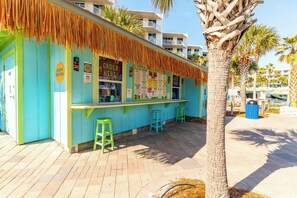 Eine Tiki-Bar mit blauer Außenfassade, einem Reetdach und grünen Hockstühlen.