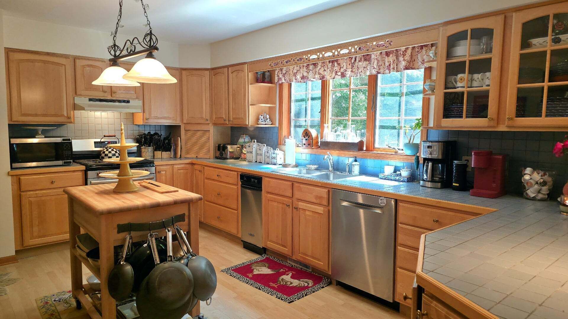A kitchen with wooden cabinets, a stainless steel dishwasher, a coffee maker, and a window with a view of greenery.