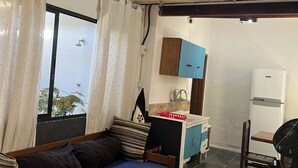 Una habitació amb un llit, una cuineta petita, un ventilador i una finestra amb cortines.