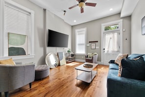 A modern living room with a flat-screen TV, a ceiling fan, and a large window with blinds. - The Benton Lounge "Historic Home Near Downtown" (St. Louis)