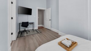 Quarto de hotel com cama, televisão fixada na parede, escrivaninha com cadeira e bandeja de madeira com toalhas.