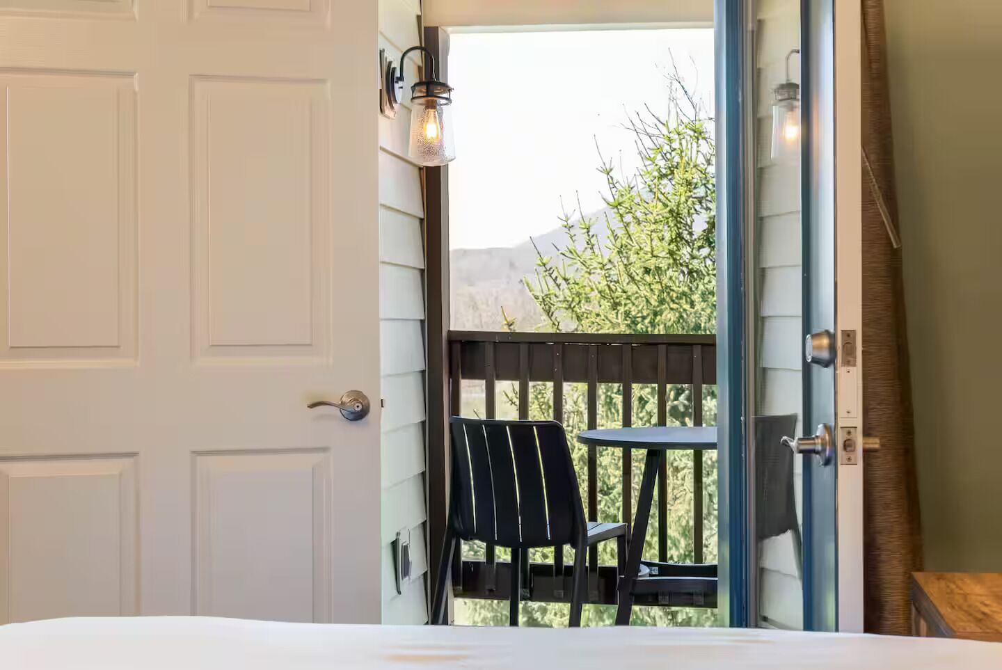 A room with a bed, a door, a balcony with a table and chairs, and a hanging light fixture.