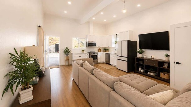 Un salon moderne avec un grand canapé d’angle beige, un téléviseur à écran plat fixé au mur et une cuisine en arrière-plan.