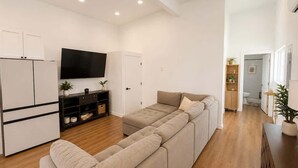 Un salon moderne comprenant un canapé, un téléviseur à écran plat et un coin cuisine avec des placards et un réfrigérateur.