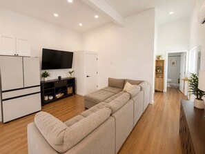 A modern living room with a sofa, a flat-screen TV, and a kitchen area with cabinets and a refrigerator. - Golden Hill Retreat with Private Hot Tub (Spring Valley)