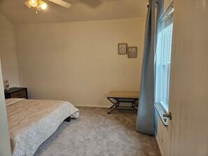 A bedroom with a bed, a bedside table, a bench, a standing lamp, and a window with curtains. - Cozy 3 Bedroom Home Near Frisco's Best Spots- Stylish&Comfy (Little Elm)