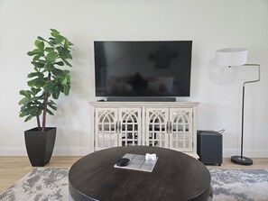 Ein Wohnzimmer mit Flachbildfernseher, einer Topfpflanze, einem runden Couchtisch und einer Stehlampe.