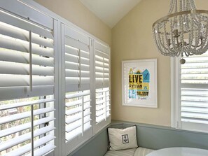 A room with white shutters, a chandelier, and a framed poster on the wall. - BlueNote by Sandpiper | Near North Beach (Seabrook Island)