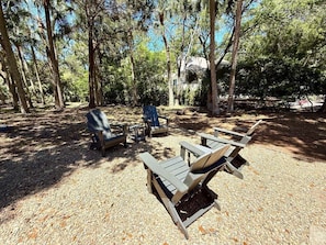 A campsite with wooden chairs and tables arranged on a gravel surface, surrounded by trees. - The Retreat by Sandpiper | Near Beach (Seabrook Island)