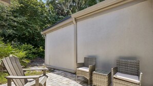 En udendørs patio med overdækket siddeområde, inklusive en træ Adirondack-stol og to fletstole med hynder.