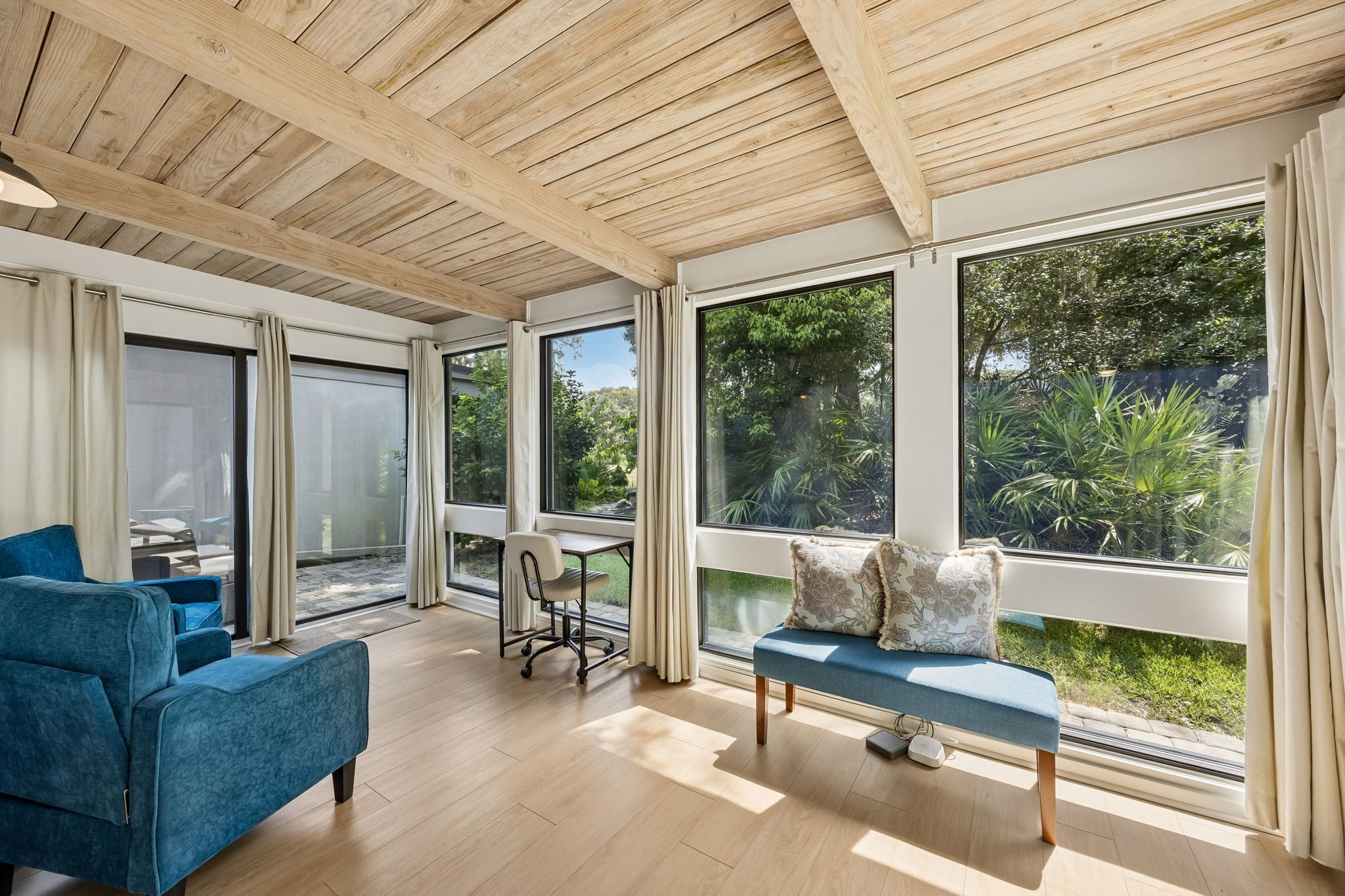 A spacious room with a wooden ceiling, large windows, a blue sofa, a dining table with chairs, and a view of greenery outside.