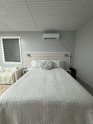 Ein Schlafzimmer mit einem Bett, einem Nachttisch, einer Lampe, einem Fenster mit Jalousien und einer Klimaanlage.