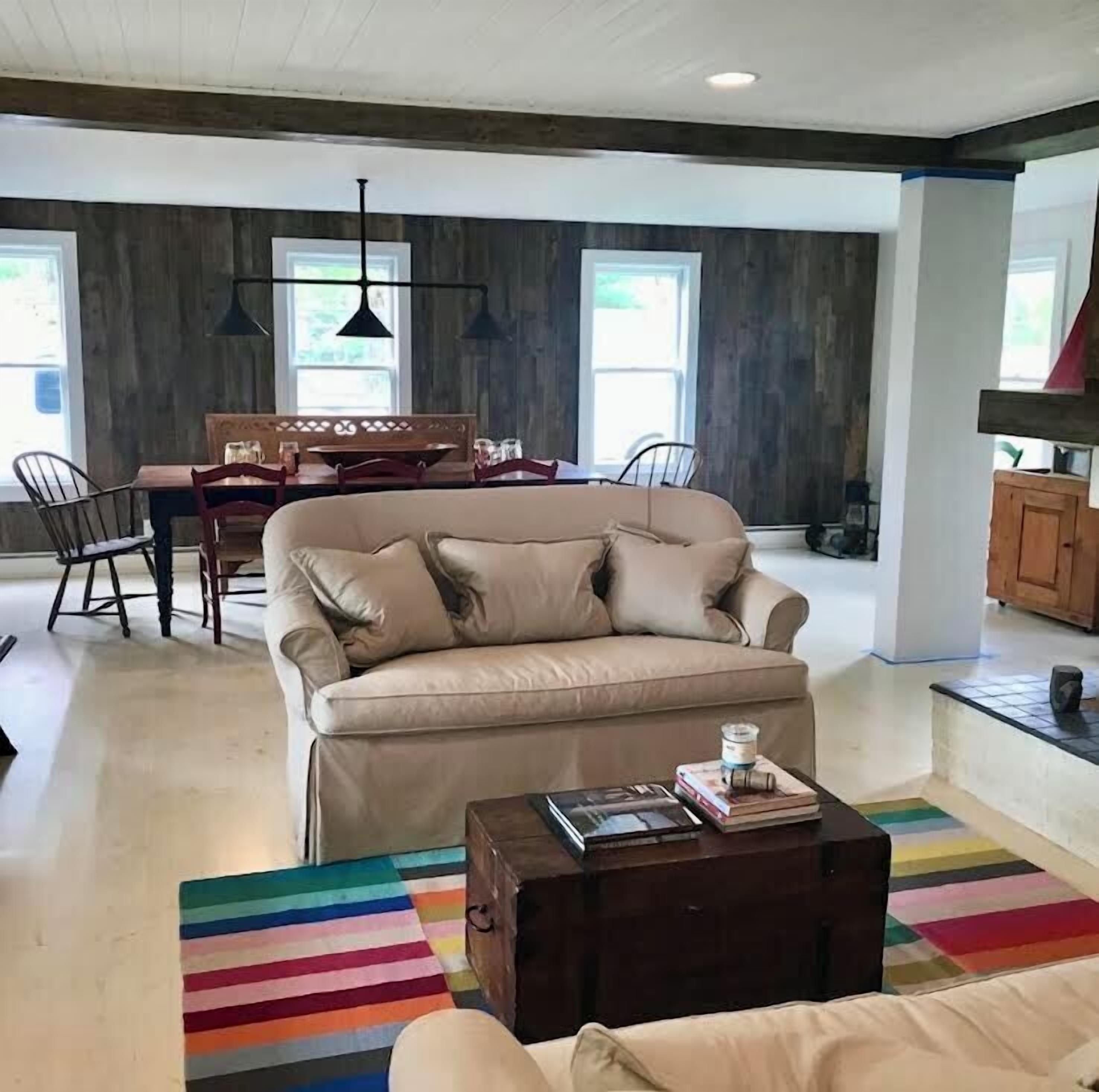 A living room with a beige sofa, a wooden coffee table, and a colorful striped rug. There is a dining area with a wooden table and chairs in the background.