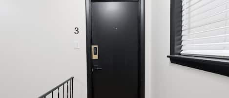 Una puerta negra con cerradura electrónica, un cartel con el número 3 encima y una ventana con persianas a la derecha.