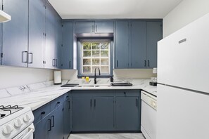 Een keuken met donkerblauwe kastjes, een witte koelkast en een raam boven de gootsteen.