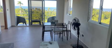 Un balcón amplio con mesa y sillas, un ventilador de pie y vista al océano.
