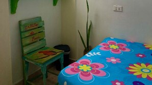 Een kleine kamer met een bed, een stoel, een groen plankje, een raam met kantgordijnen en een hanglamp.