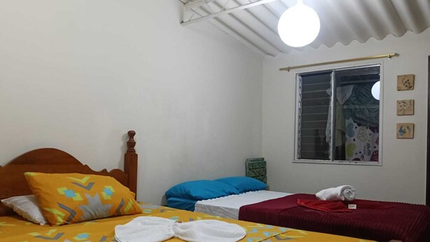 A room with two beds, one with a yellow patterned bedspread and the other with a red one. There are towels on the beds, and a window with curtains. - Room-room Mirador, Casa Del Pueblo (Jardin)