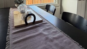 Un comedor con una mesa dispuesta para dos, una ventana que ofrece vista a una cerca de madera, y una cocina visible al fondo.
