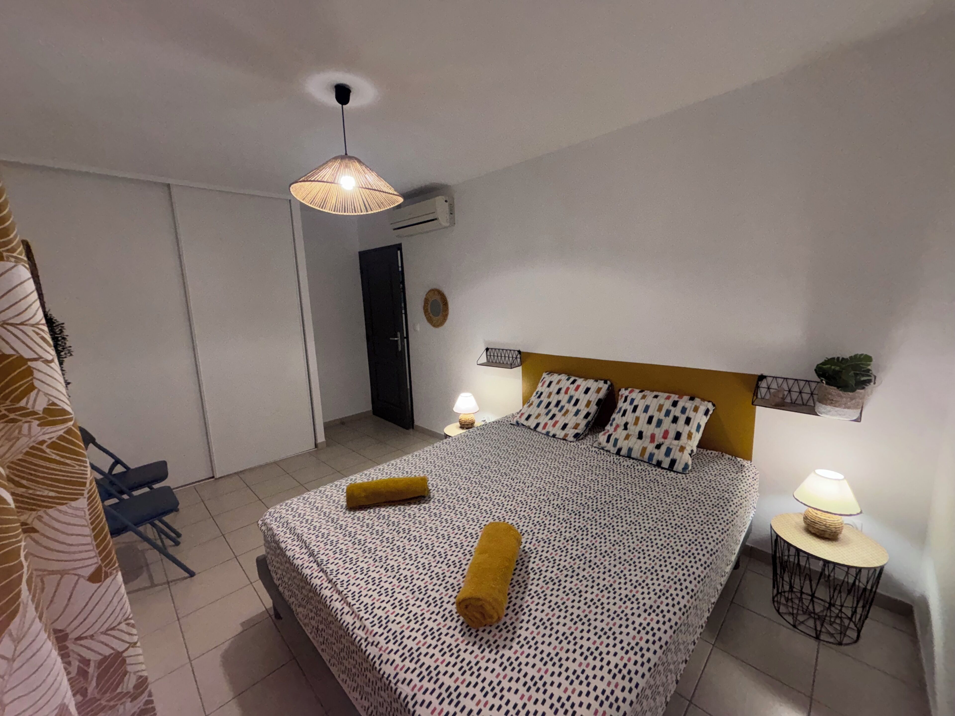 A bedroom with a bed, bedside lamps, a hanging light fixture, a door, and a small table with a lamp.