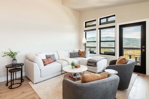 A modern living room with a sectional sofa, a round coffee table, and two armchairs. There is a small side table with a plant, and the room has large windows with a view of the countryside. - Min to Deer Valley East Newest Terrain, Community Amenities, Free Ski Shuttle (Heber City)