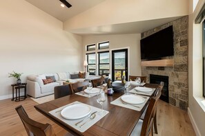 Un coin repas avec une longue table en bois dressée pour quatre personnes, une cheminée en pierre et un canapé en arrière-plan.
