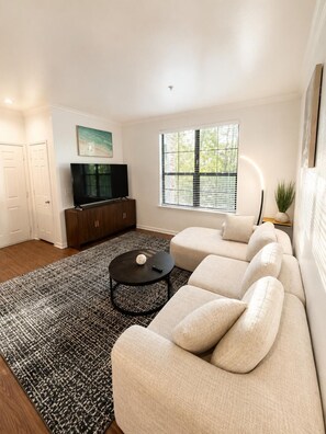 A modern living room with a sectional sofa, a flat-screen TV, and a patterned rug. - Stylish 1BR Resort Retreat Near The Woodlands (Conroe)