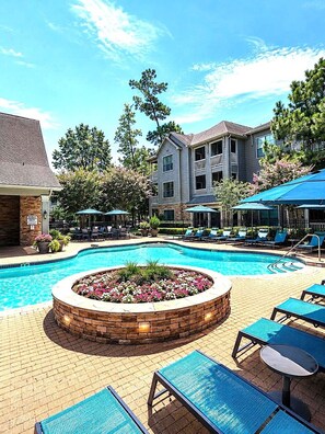 A swimming pool with lounge chairs and umbrellas, surrounded by a brick patio and a building with balconies. - Stylish 1BR Resort Retreat Near The Woodlands (Conroe)