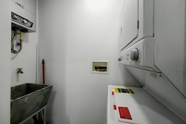 A narrow hallway with a white wall, a metal bucket, a red-handled tool, and a white appliance with a red label.
