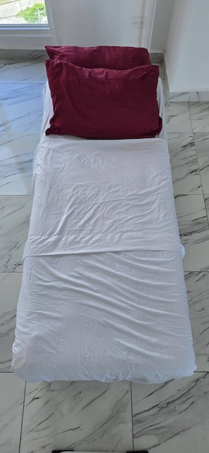 Un lit avec une literie blanche et un seul oreiller rouge, dans une pièce dont le sol est recouvert de marbre.