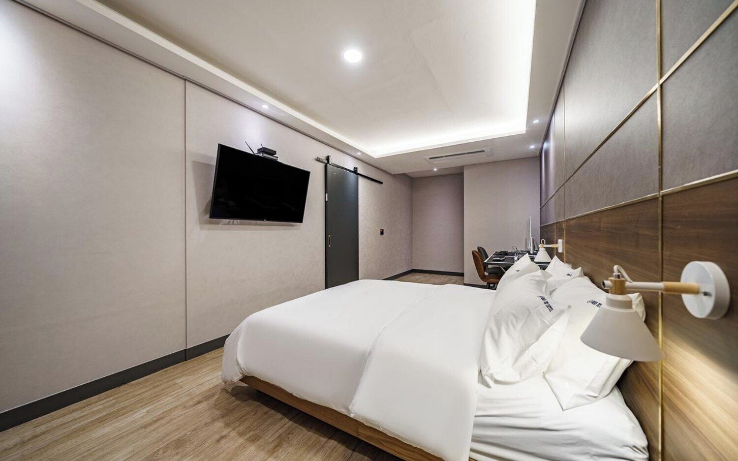 A hotel room with a bed, a TV mounted on the wall, a desk with a chair, and a bedside table with a lamp.