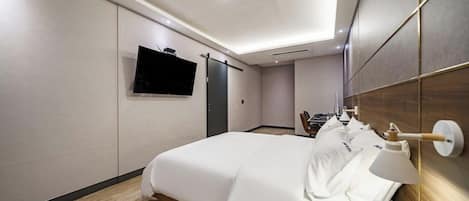 Habitación de hotel con cama, televisor montado en la pared, escritorio con silla y mesita de noche con lámpara.