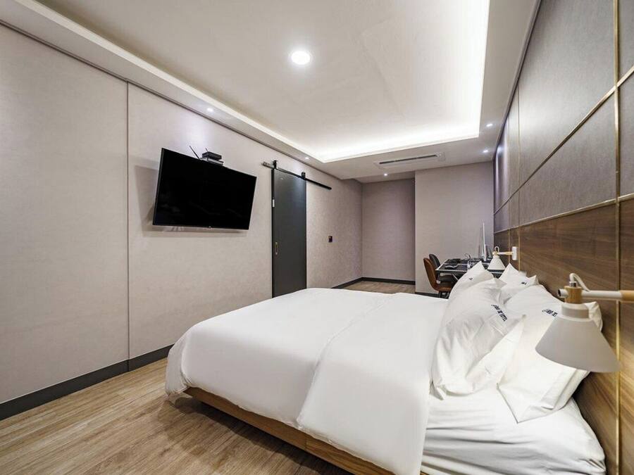 A hotel room with a bed, a TV mounted on the wall, a desk with a chair, and a bedside table with a lamp.