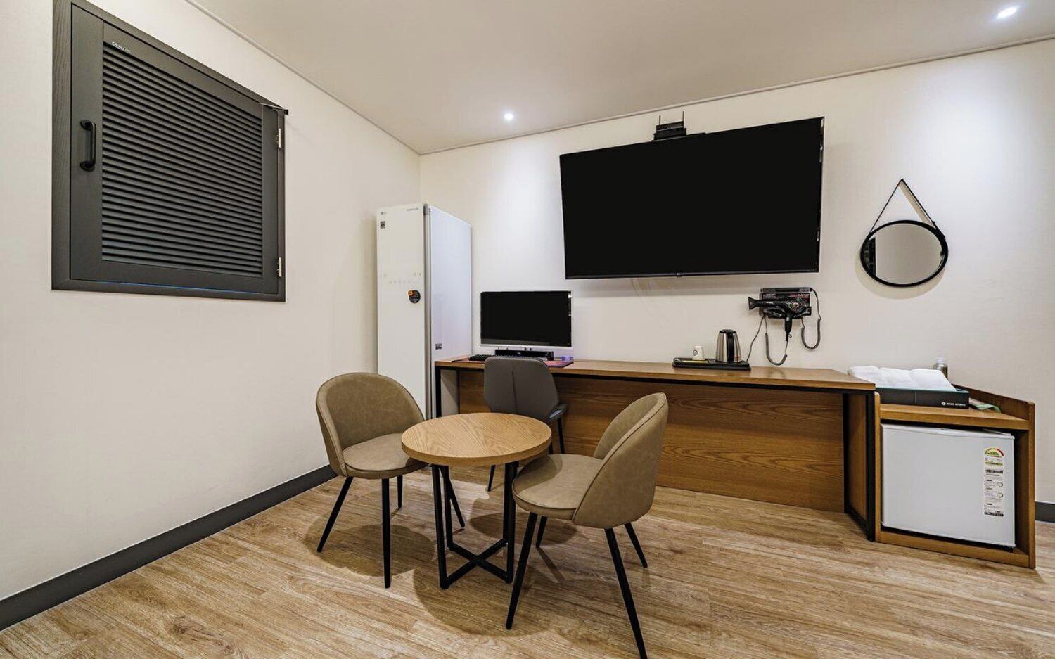 A compact hotel room with a kitchenette, a flat-screen TV mounted on the wall, a small refrigerator, a desk with a computer, and two chairs around a small round table.