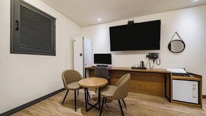 Habitación de hotel compacta con kitchenette, un televisor de pantalla plana en la pared, un refrigerador pequeño, un escritorio con computadora y dos sillas alrededor de una mesita redonda.