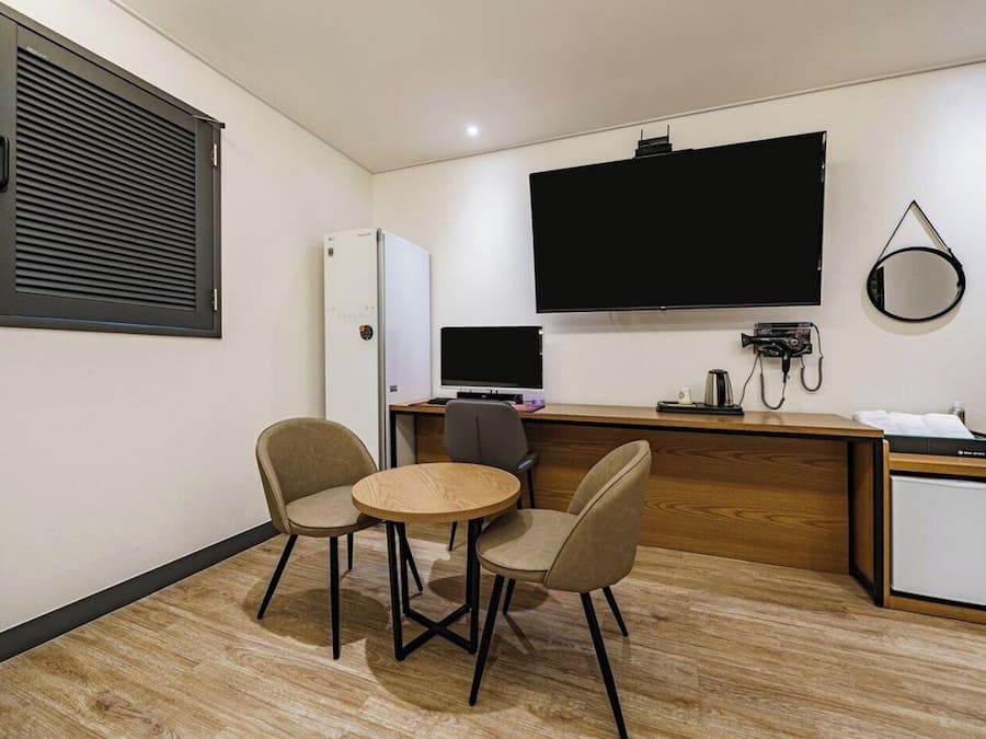 A compact hotel room with a kitchenette, a flat-screen TV mounted on the wall, a small refrigerator, a desk with a computer, and two chairs around a small round table.