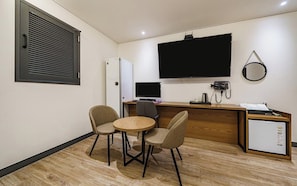 A compact hotel room with a kitchenette, a flat-screen TV mounted on the wall, a small refrigerator, a desk with a computer, and two chairs around a small round table.
